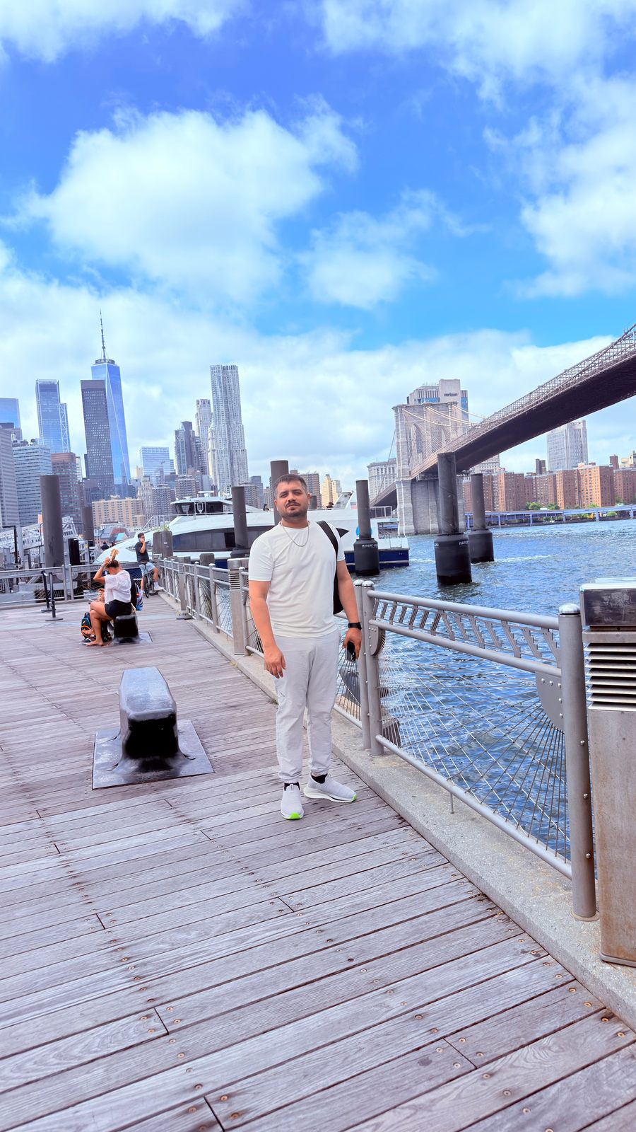 Wali Zuberi on a waterfront boardwalk with the Brooklyn Bridge and the Lower Manhattan skyline behind him